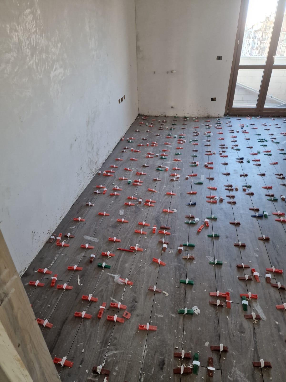 Posa di pavimento in gres effetto legno in una stanza al grezzo, con distanziatori livellatori rossi e verdi e pareti intonacate.