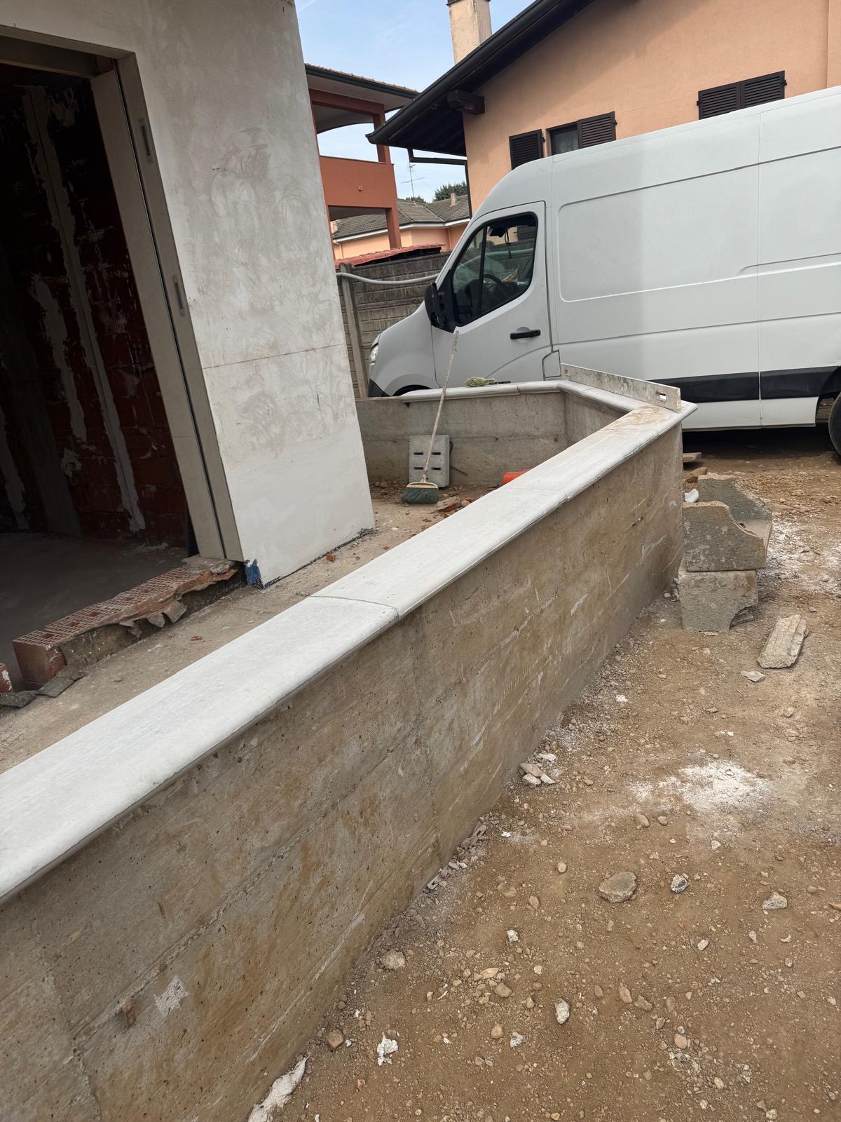 Muretto esterno in cemento con copertina lungo l'ingresso di un edificio in costruzione; cortile in terra e furgone da cantiere.